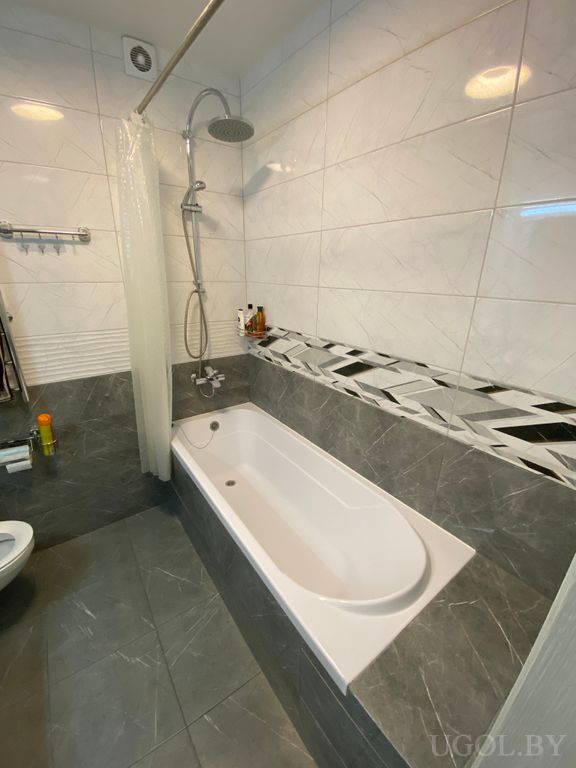 Интерьер с белой ванной, серой плиткой, декоративной вставкой и шторкой с геометричным узором.