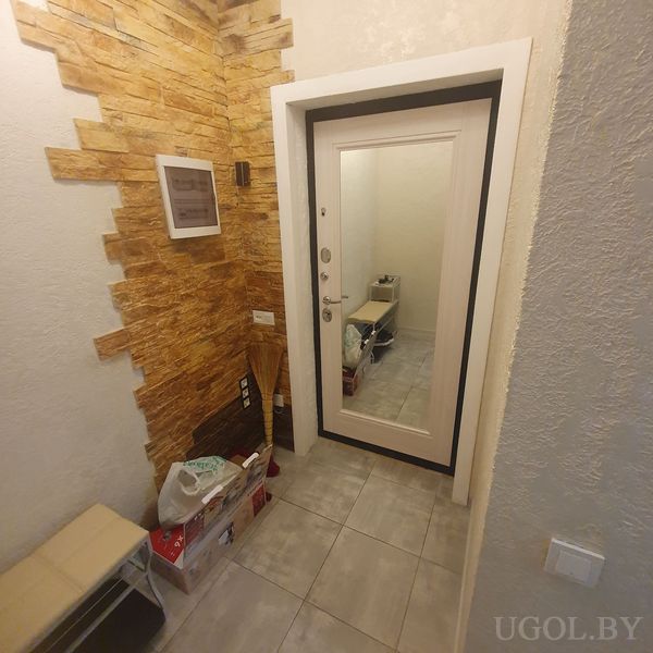 Узкий коридор с белой дверью, декоративной каменной стеной, светильником и предметами хранения.