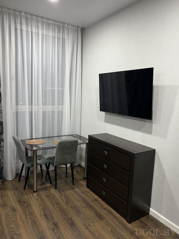 Минималистичный угол с стеклянным столом, двумя серыми стульями, тумбой с телевизором и белыми шторами.