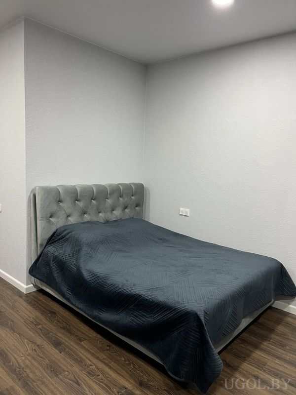 Спальня с серым изголовьем, тёмным покрывалом, серыми стенами и розеткой у кровати на фоне тёмного пола.