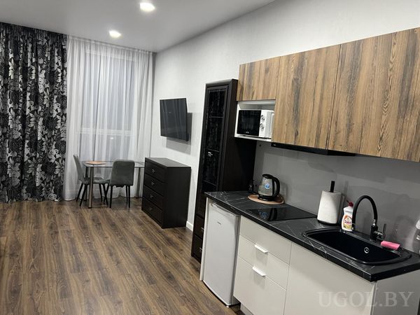 Компактная кухня с чёрной мойкой, белыми и деревянными фасадами, обеденной зоной и телевизором на тумбе.