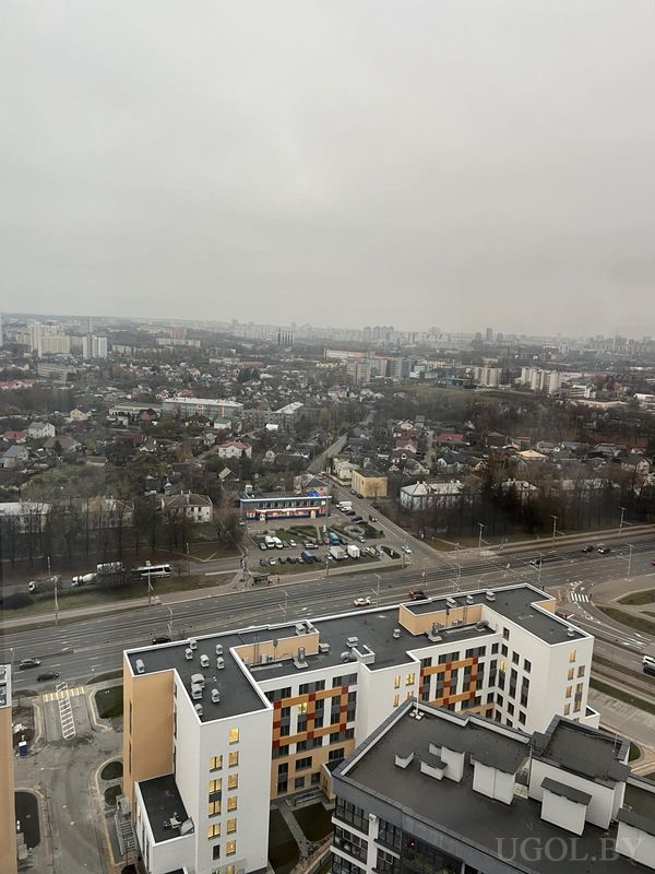 Панорама с высоты на жилые дома, дорогу с машинами, торговый центр и застройку на фоне серого неба.