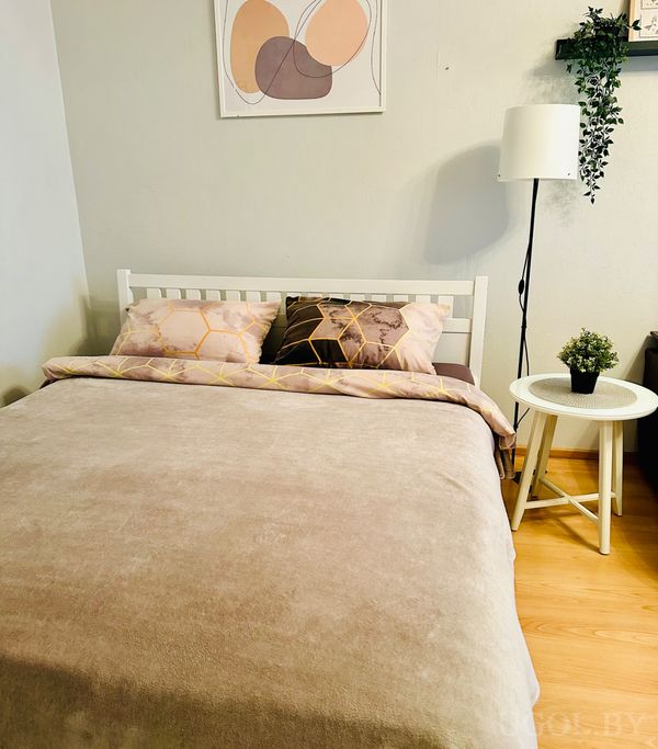 Комната с кроватью, бежевым покрывалом, подушками с геометричным узором, торшером и растением на белом столике.