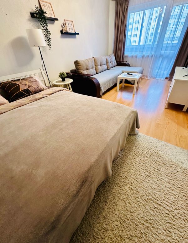 Жилая зона с кроватью, диваном, журнальным столиком, телевизором, полками с декором и окнами с тюлем и шторами.