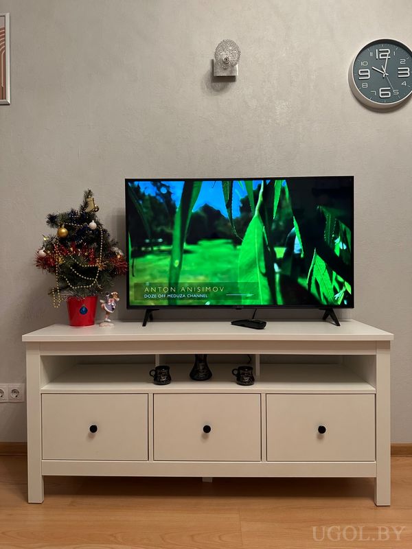 Интерьер с телевизором на белой тумбе, новогодним декором, часами на стене и тремя кружками на полке.