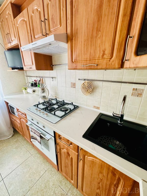 Кухонная зона с плитой, вытяжкой, мойкой, чайником, телевизором и фартуком с бежевым узором.