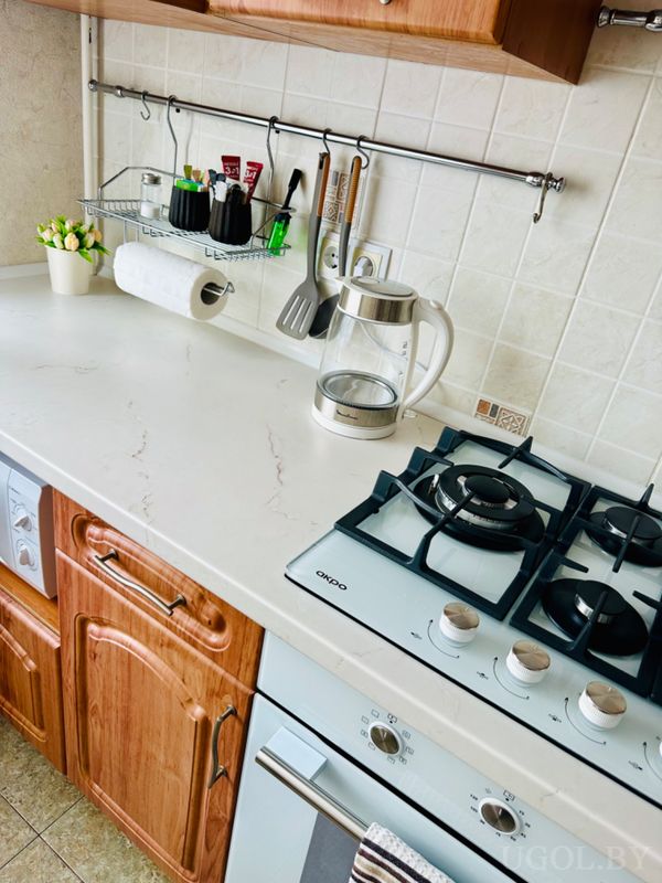 Рабочая зона с плитой, чайником, рейлингом с приборами, полкой с чашками и растением на фоне светлой плитки.