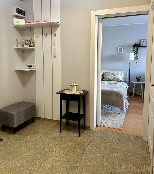 Интерьер с чёрным столиком, полкой с декором, серой банкеткой и видом на спальню с серым текстилем и лампой.