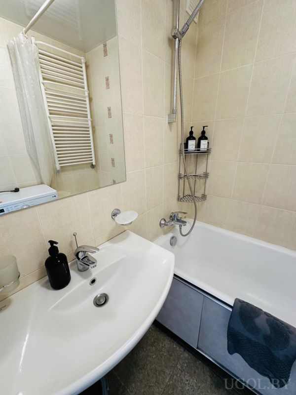 Комната с умывальником, зеркалом, ванной, чёрным дозатором, стеклянной ёмкостью и плиткой в бежевых тонах.