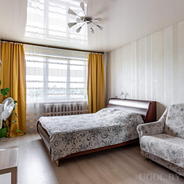 Кровать с цветочным покрывалом, мягкое кресло, горшечное растение, вентилятор и окно с жёлтыми портьерами.