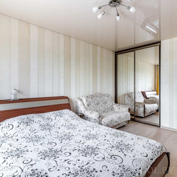 Кровать с чёрно-белым покрывалом, кресло, зеркальный шкаф, светлый пол и потолочный светильник.