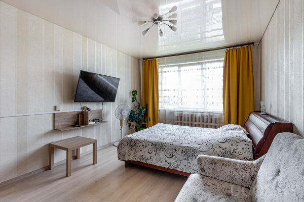 Кровать, кресло, телевизор на стене, полка с вентилятором и окно с яркими шторами на фоне светлых обоев.