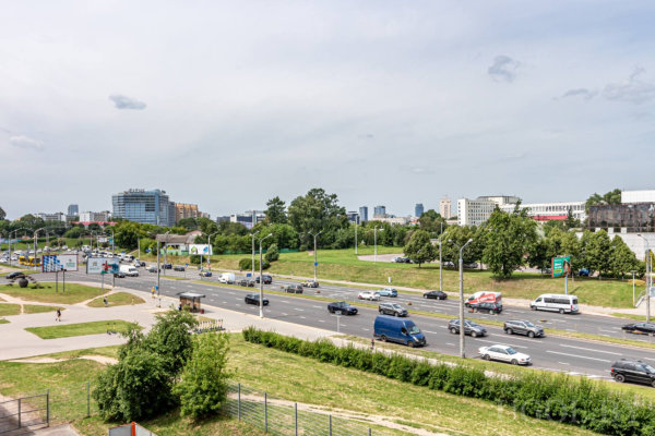 Многополосная дорога, машины, автобусы, остановка, фонари, зелёные зоны и современные здания на фоне.