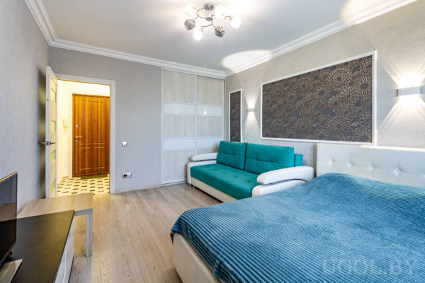 Кровать с бирюзовым пледом, диван, ТВ-зона, светлый пол и декоративная панель с цветочным узором над диваном.