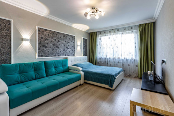 Бирюзовый диван, кровать с синим покрывалом, три чёрно-белых панно, телевизор на тумбе и окно с зелёными шторами.