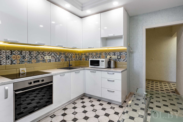 Глянцевые белые шкафы, варочная панель, микроволновка, плитка с жёлто-чёрным узором и стеклянный стол.