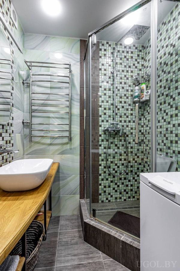 Раковина на деревянной столешнице, стеклянная душевая с мозаикой, полотенцесушитель и стиральная машина.