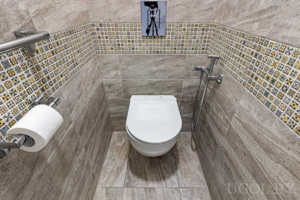 Навесной унитаз, серо-бежевые стены, розетка, гигиенический душ и декоративная плитка с изображением камеры.