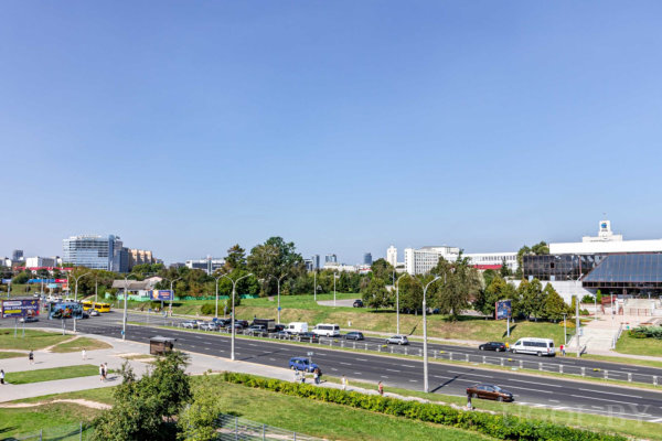 Широкая дорога с машинами и автобусом, пешеходы, зелёные зоны, современные здания и ясное небо.