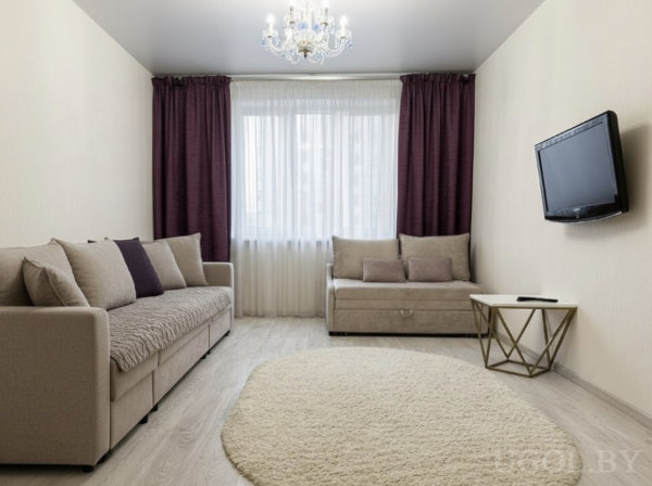 Два бежевых дивана напротив друг друга, круглый светлый ковёр, телевизор на стене, золотой столик и большое окно с тюлем и фиолетовыми шторами.