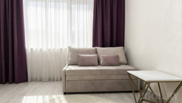 Бежевый диван с подушками, рядом белый столик с золотым каркасом, большое окно с тюлем и фиолетовыми шторами, светлый деревянный пол.