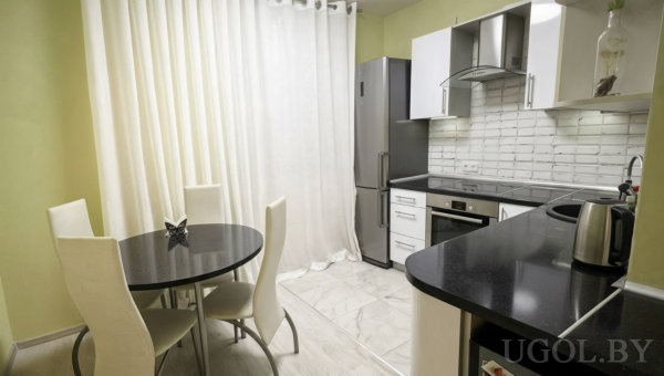 Белая кухня с чёрной столешницей, круглым чёрным столом и белыми стульями, на фоне холодильник и духовка, окно с тюлем.