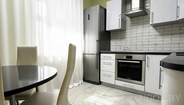 Современная кухня с белыми фасадами, тёмной столешницей, круглым столом и мягкими стульями, светлый пол и тюль на окне.