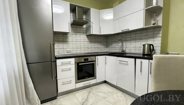 Белые шкафы, чёрная столешница, варочная панель, раковина и холодильник, светлый пол с мраморным рисунком и тюль у окна.