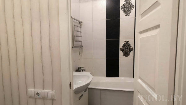 Белая ванна и раковина, вертикальная полоса чёрной плитки с узором, металлическая полка и светлая дверь с панелями.