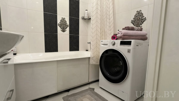 Белая ванна с чёрными декоративными вставками на стене, рядом стиральная машина с аккуратно сложенными полотенцами и светлая шторка.