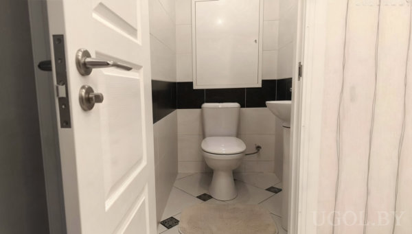 Белый унитаз и раковина, стены с горизонтальной чёрной полосой, светлый пол с чёрными акцентами и бежевый коврик.