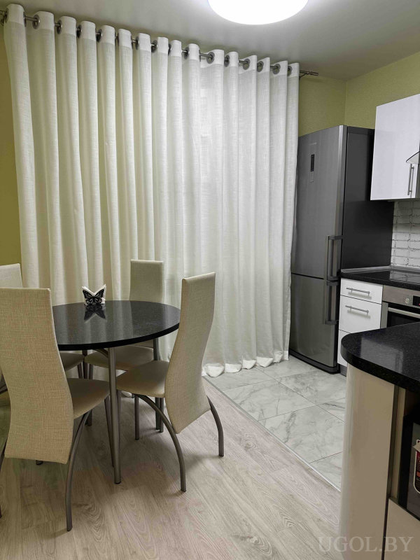 Интерьер с чёрным столом, бежевыми стульями, белыми шторами, холодильником, плитой и светлым зонированным полом.