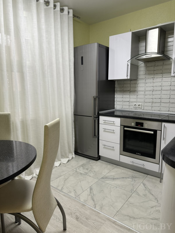 Рабочая зона с плитой, вытяжкой, холодильником, чёрной столешницей, белыми шкафами и мраморной плиткой.