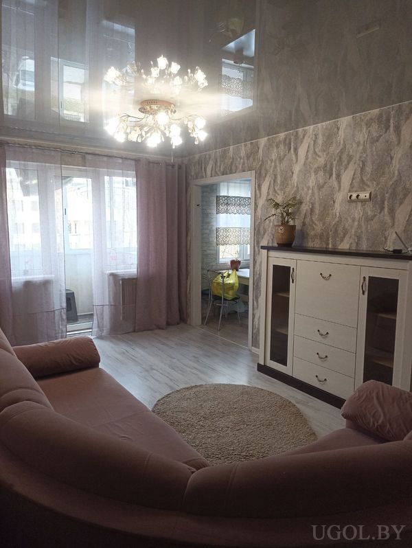 Комната с изогнутым диваном, круглым ковром, белой тумбой, люстрой и выходом в столовую зону с жалюзи.