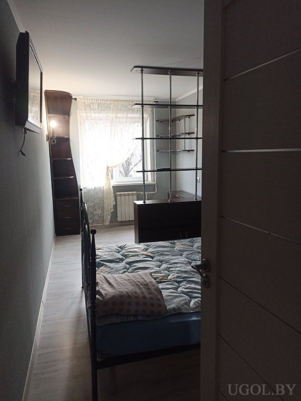 Компактная комната с кроватью, телевизором на стене, открытыми полками и окном с белыми шторами.