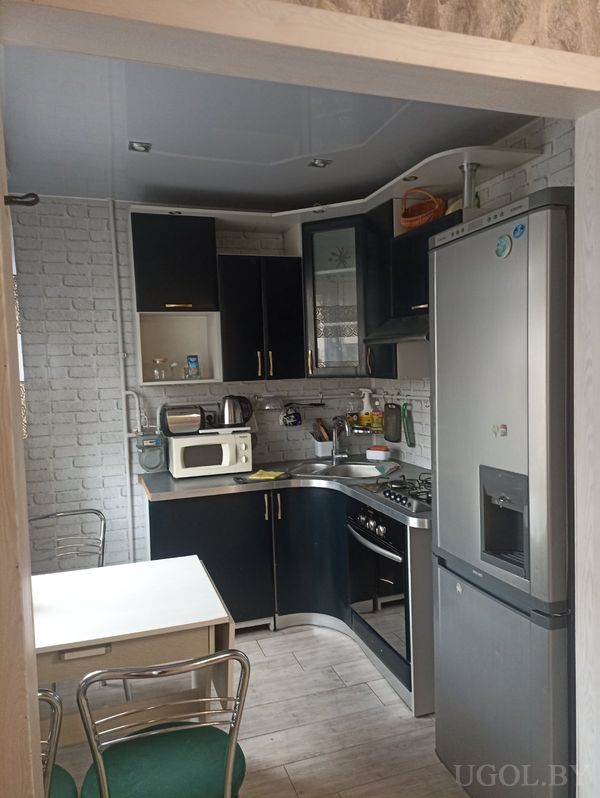 Угловая кухня с чёрными шкафами, плитой, вытяжкой, холодильником с диспенсером и столом с зелёными сиденьями.