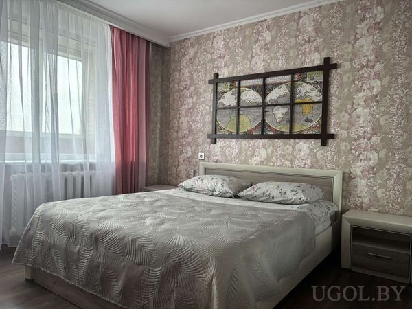 Уютная спальня с кроватью в светлом текстиле, настенной композицией из карт и розовыми шторами на фоне цветочных обоев.