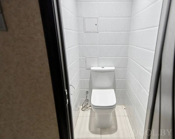 Узкий туалет с белой плиткой, гигиеническим душем, люками и светлым полом.