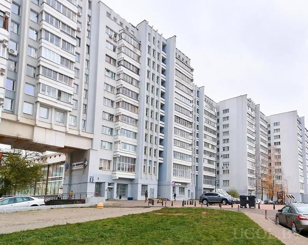 Ряд многоэтажек с магазинами на первом этаже, парковкой и газоном на фоне пасмурного неба.