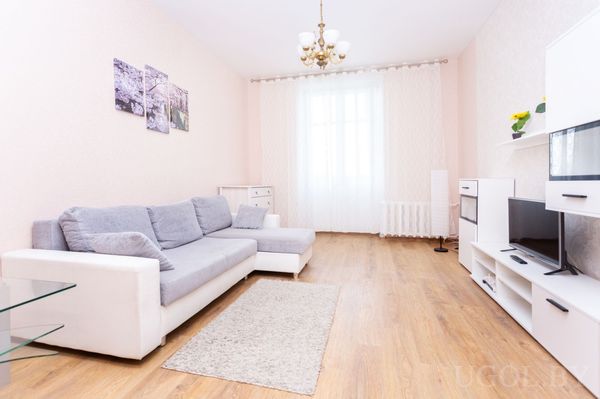 Современная гостиная с серо‑белым диваном, телевизором на белой тумбе, люстрой и большим окном с белыми шторами.