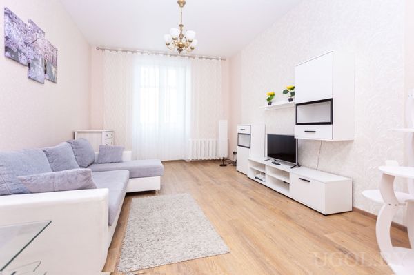 Светлая гостиная с серым диваном, телевизором на белой тумбе, обеденным столом и большим окном с белыми шторами.