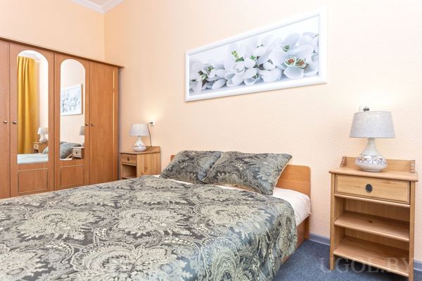 Спальня с кроватью в зелёном покрывале, прикроватными тумбами и лампами, шкафом с зеркальными дверцами и картиной с цветами.