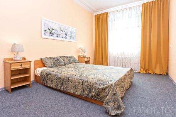 Спальня с кроватью, прикроватными тумбами и лампами, серым ковролином и большими окнами с белыми и жёлтыми шторами.