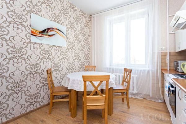 Светлая кухня с деревянным столом, белыми шкафами, плитой и абстрактной картиной на стене.