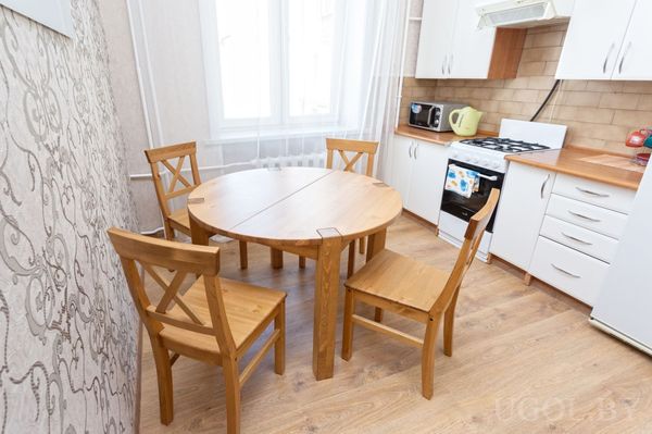 Компактная кухня с белыми шкафами, плитой, круглым деревянным столом и окнами с белыми шторами.