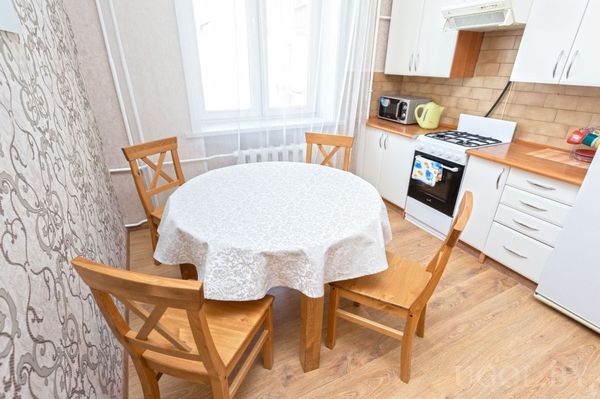 Кухня с белыми шкафами, плитой, круглым столом с белой скатертью и окнами с белыми шторами.