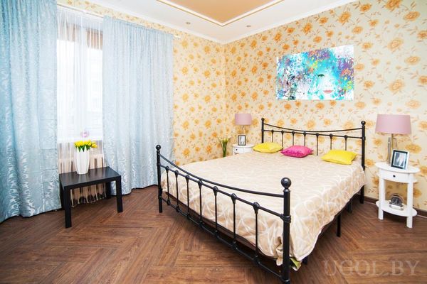 Уютная спальня с металлической кроватью, бежевым покрывалом, яркими подушками и абстрактной картиной над изголовьем.