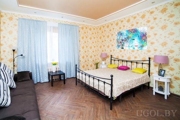 Комната с металлической кроватью, диваном с подушками, абстрактной картиной и столиком с вазой жёлтых цветов у окна.