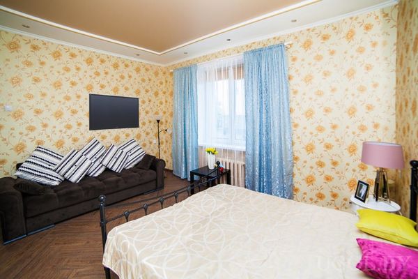 Комната с кроватью и яркими подушками, диваном с полосатыми подушками, телевизором на стене и столиком с лампой у окна.