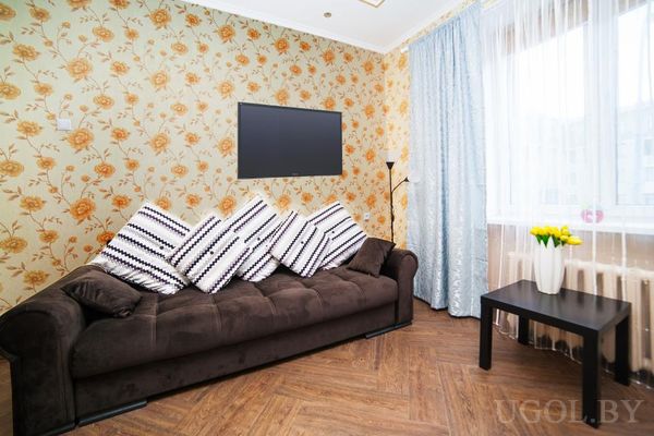 Уютный уголок гостиной с тёмным диваном, полосатыми подушками, телевизором на стене и столиком с вазой жёлтых тюльпанов.